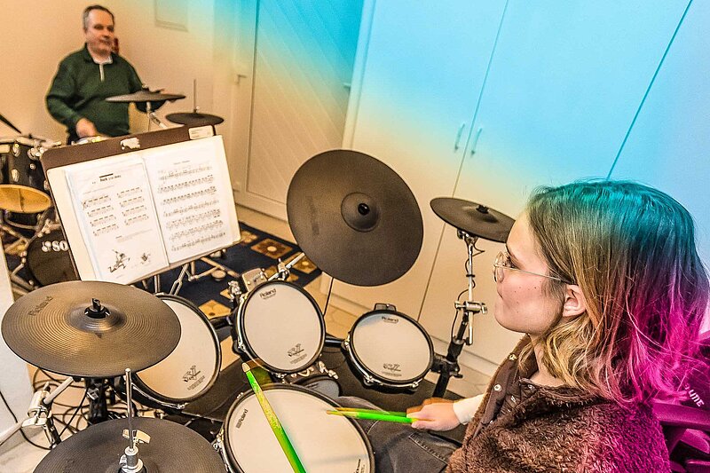 Jugendliche übt konzentriert an einem E-Drumset; Im Hintergrund der Lehrer an einem eigenen Drumset